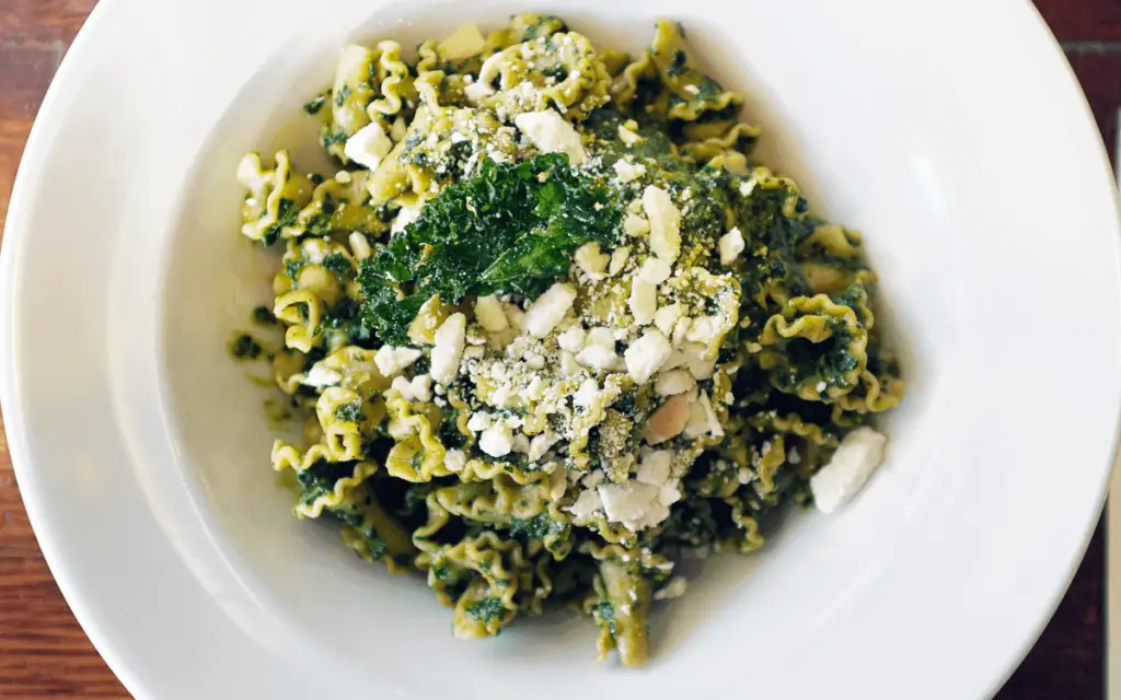 A plate of Mafaldine pasta generously coated with zesty pea shoot and Meyer lemon pesto, garnished with fresh pea shoots and a sprinkle of lemon zest, set against a rustic table backdrop.