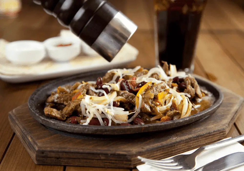 Hand sprinkling homemade fajita seasoning over a sizzling meat dish, highlighting the rich blend of spices.