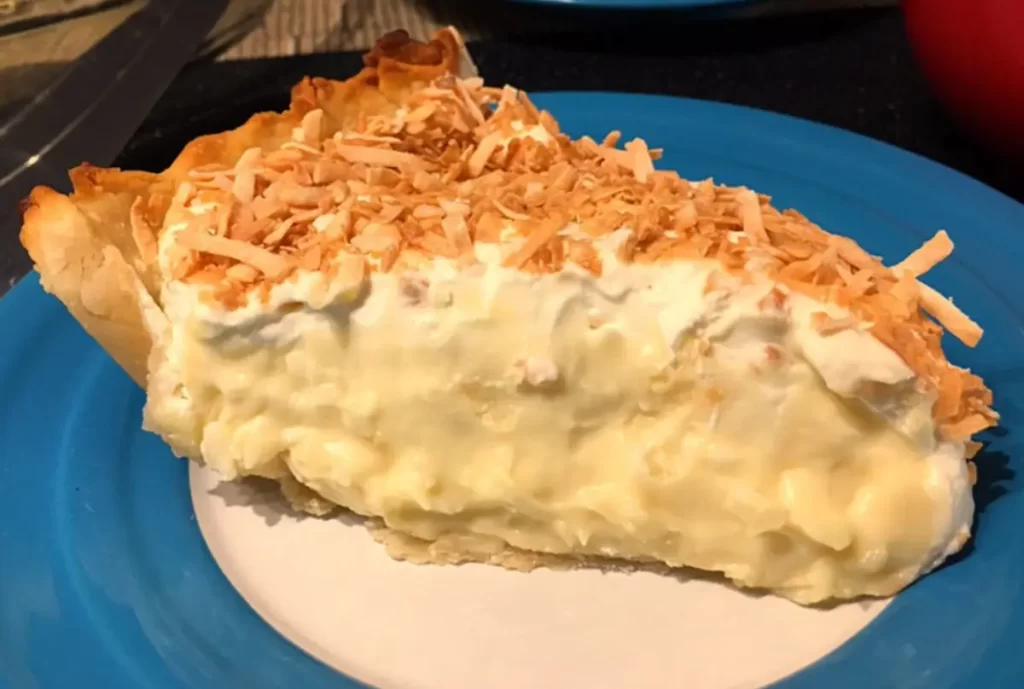 A delectable slice of Coconut Cream Pie, featuring a golden crust filled with creamy coconut custard, topped with a generous layer of fluffy whipped cream and garnished with toasted coconut flakes.