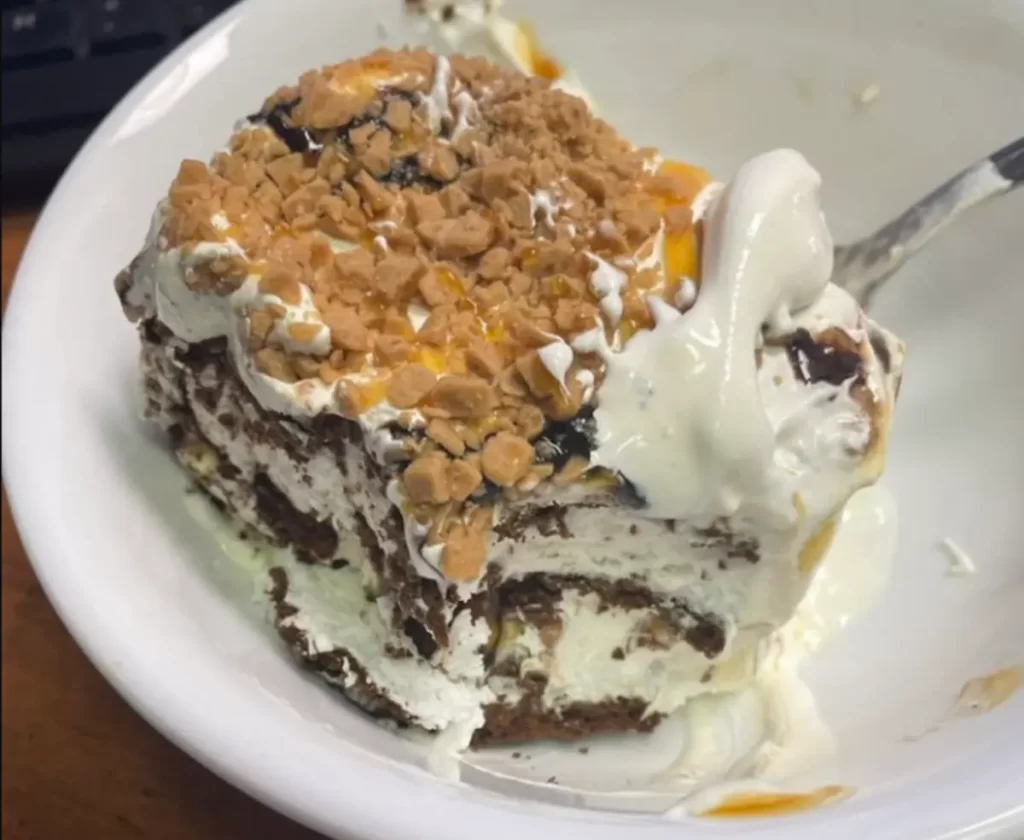 Layered Ice Cream Sandwich Cake with cream cheese mixture, topped with whipped cream, chocolate chips, and caramel drizzle on a serving plate.