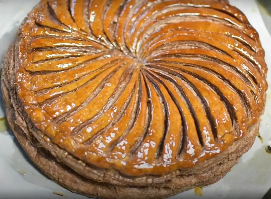 A golden-brown Hazelnut-Praline King's Cake with a glossy finish, made with rich chocolate puff pastry, garnished with a decorative score pattern on top, symbolizing the traditional Epiphany celebration.