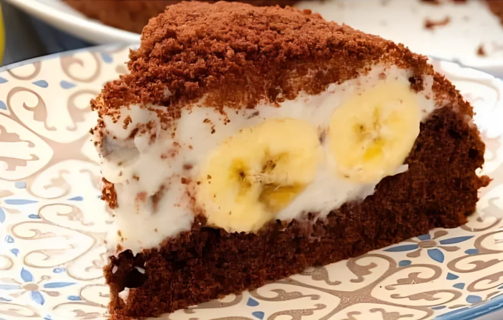 A delectable slice of Banana Dome Cake showcasing layers of moist chocolate cake, fresh banana slices, and creamy whipped cream topped with fine crumbs, served on a plate.