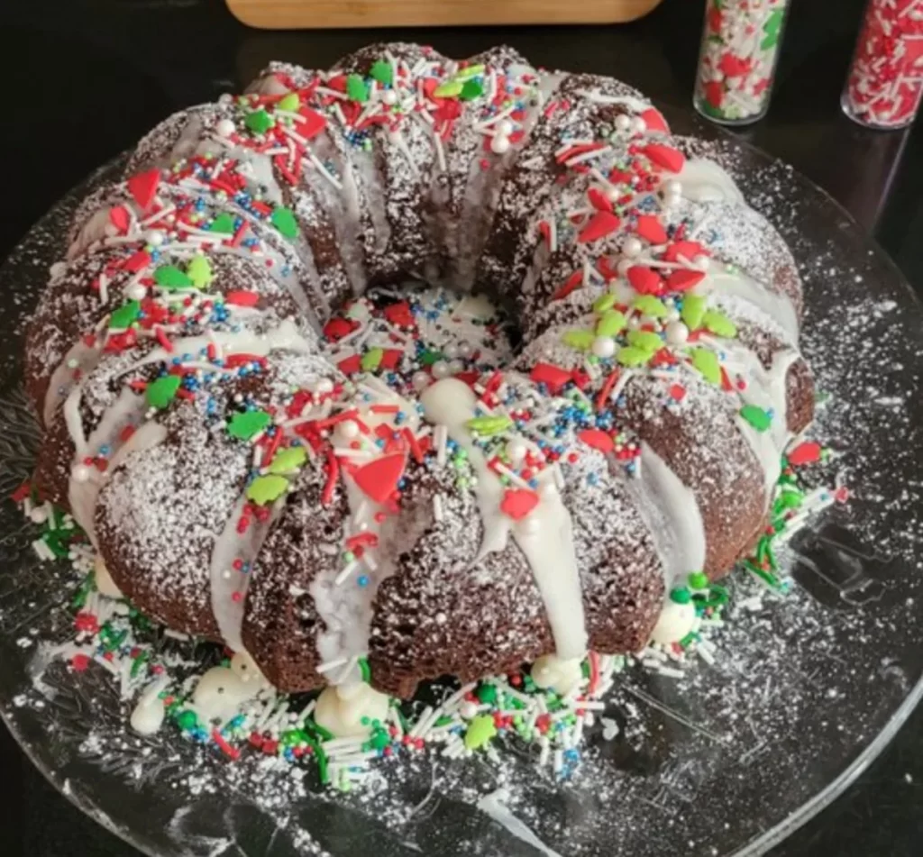 A chocolate Bundt cake with white icing and colorful holiday sprinkles on a glass plate, with a custom cutting board in the background.