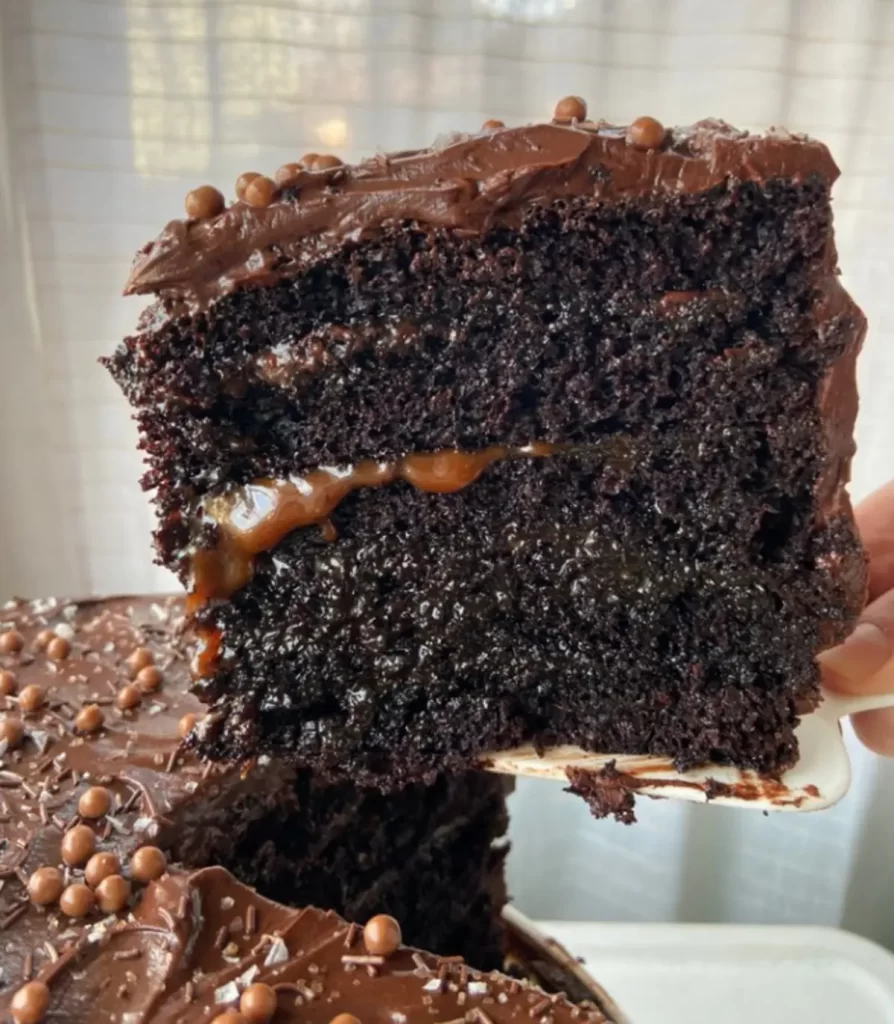 A slice of moist chocolate layer cake with rich caramel filling and glossy chocolate frosting, topped with delicate chocolate pearls.