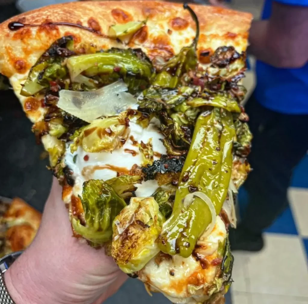 A slice of pizza with roasted Brussels sprouts and blistered shishito peppers on top, showcasing a unique and savory topping combination.