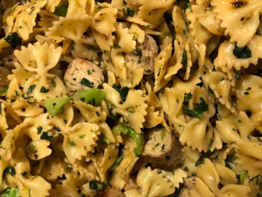 A plate of bow tie pasta with sautéed greens and sliced vegan sausage in a garlic olive oil dressing.