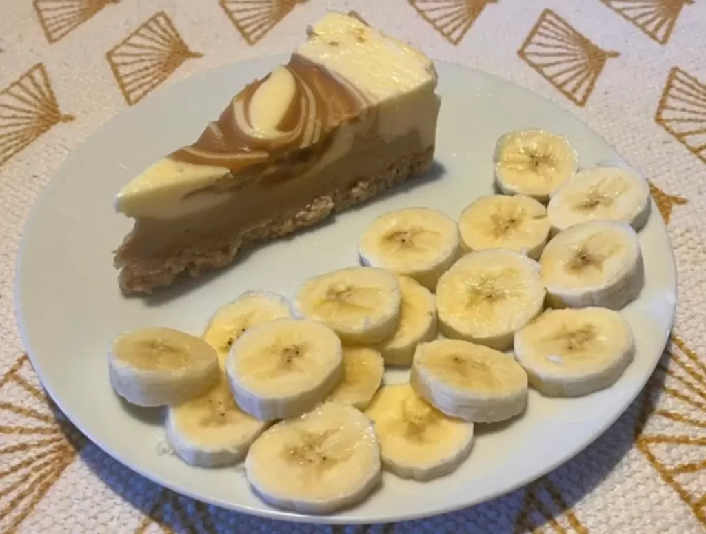 A slice of vanilla and caramel cheesecake with banana slices on a white plate.