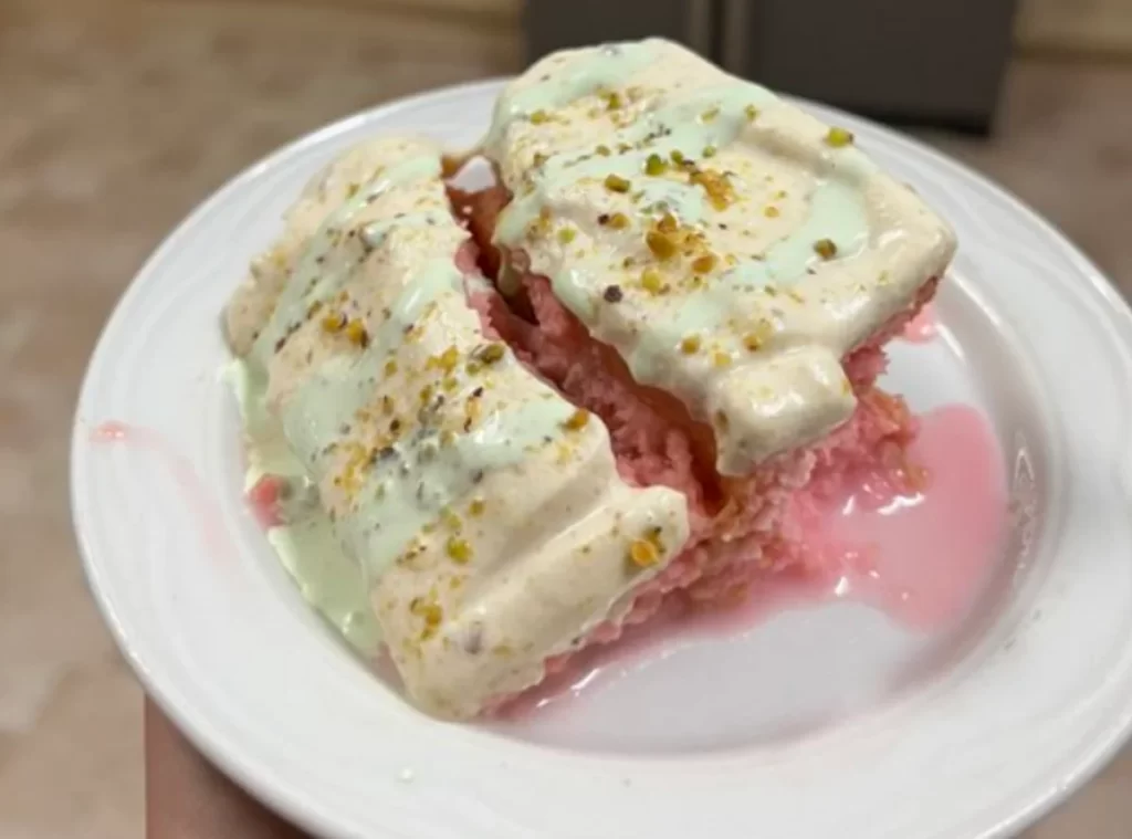 A delectable slice of Rose Milk Cake, showcasing its soft and spongy texture, elegantly topped with a creamy layer of frosting, sprinkled with chopped pistachios and adorned with delicate dry rose petals, all served on a pristine white plate.